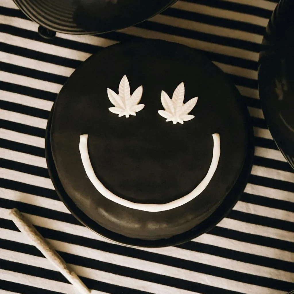 A chocolate cake on a striped table with frosting layed out in a smiley face and cannabis plants for eyes